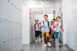 Diverse group of children with backpacks running laughing down a hallway lined with lockers