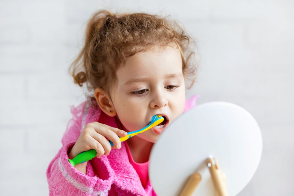 Little girl in pink robe brushing teeth with colorful toothbrush